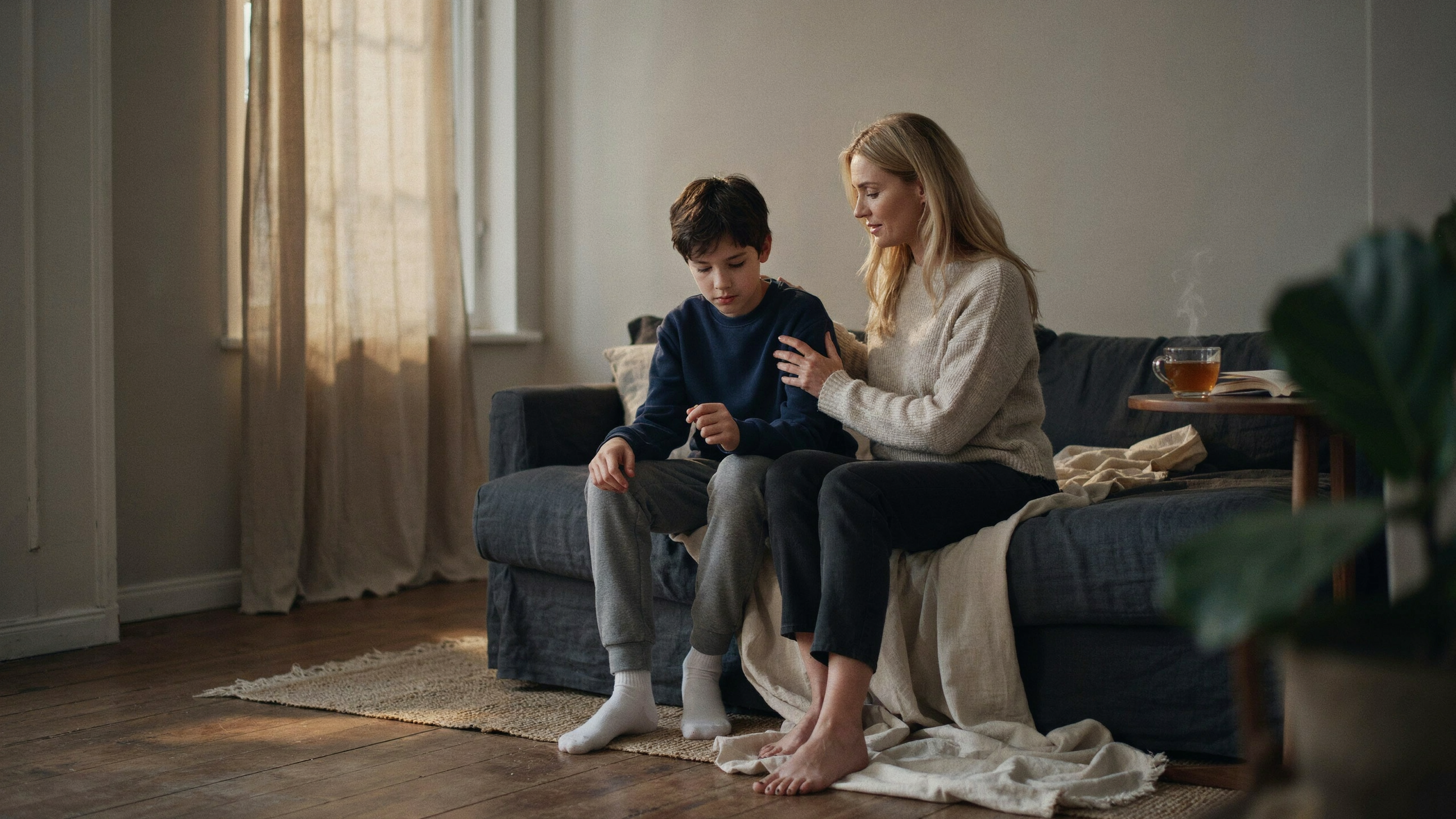 A mother sits close beside her young son in warm golden window light, one hand resting on him, comforting and encouraging him after a hard day