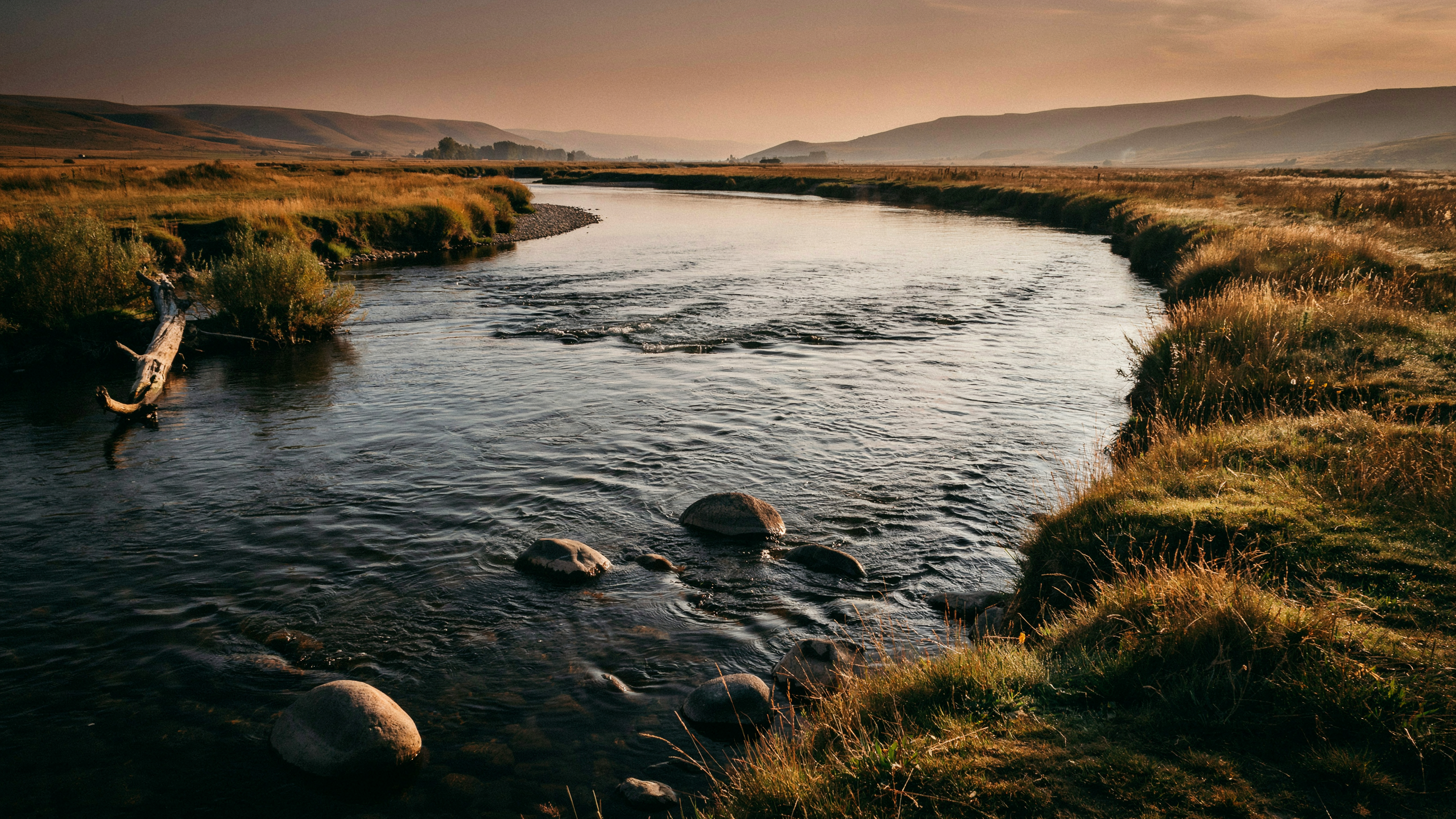 A wide river bending gently through open landscape at dawn, warm copper light on moving water with visible ripples over stones in the current, distant misty hills, dry grasses lining the banks
