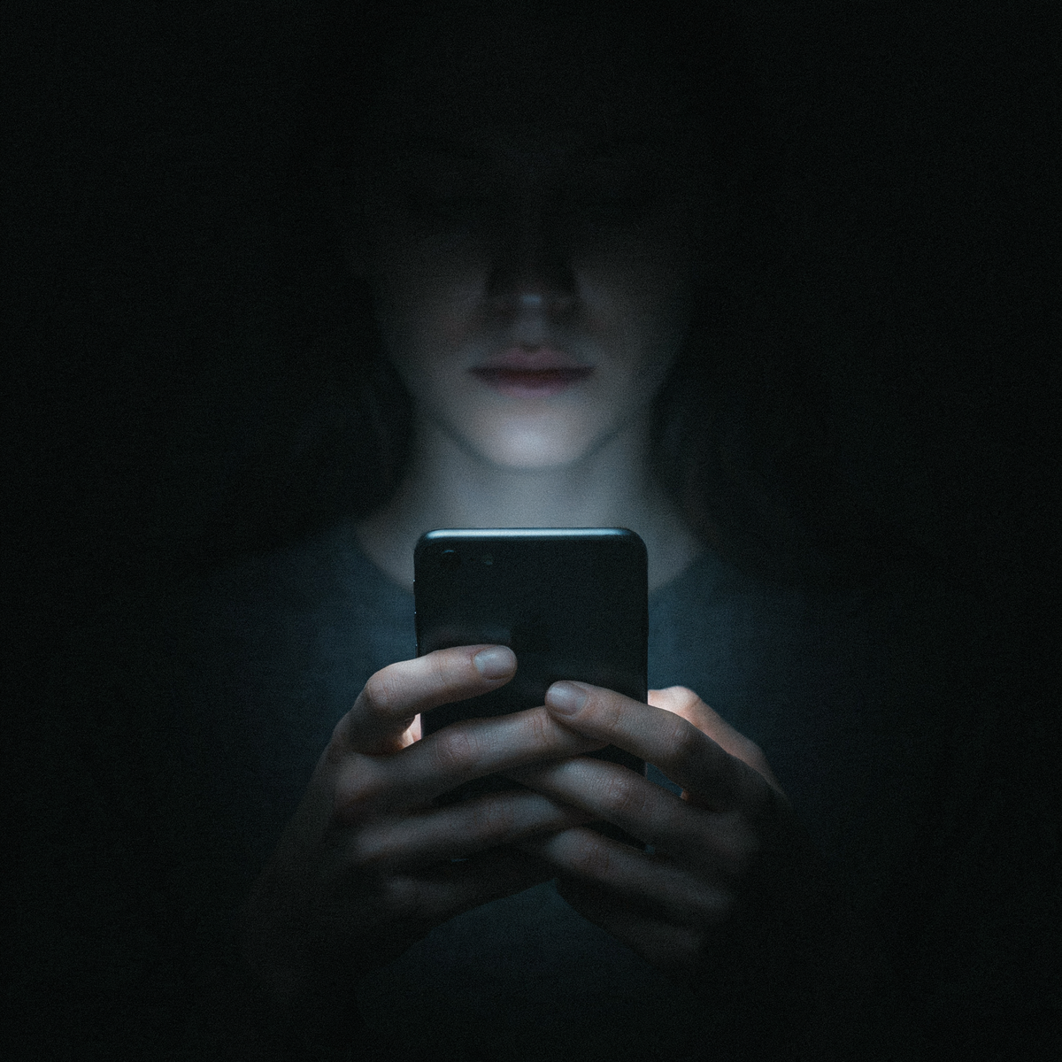 A single hand holding a smartphone at night, the screen's cold blue-white glow lighting the underside of the hand and faintly underlighting a chin, deep black surroundings, the phone as the only light source in the room