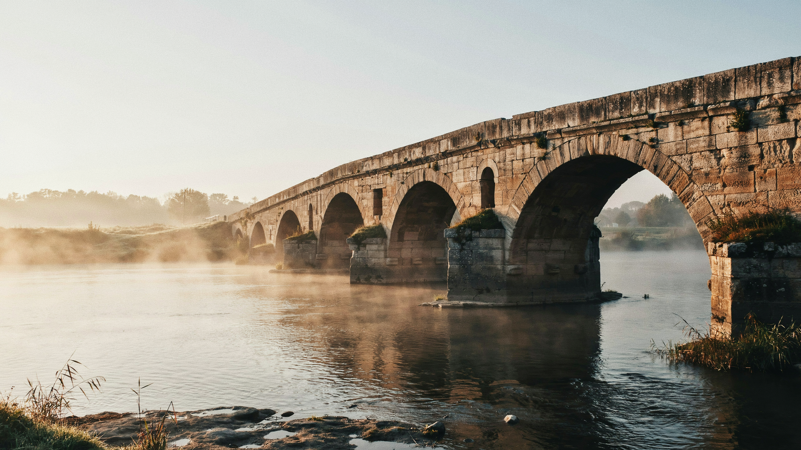 An ancient stone arch bridge glowing in golden dawn light over a calm misty river, still standing and still in use after centuries
